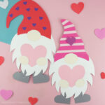 Two Valentine paper gnome crafts laying side by side on a pink background with heart stickers scattered around.