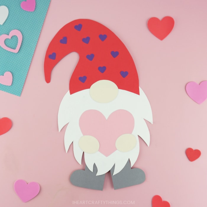 Valentine paper gnome with a red floppy hat decorated with purple hearts holding a large pink heart.