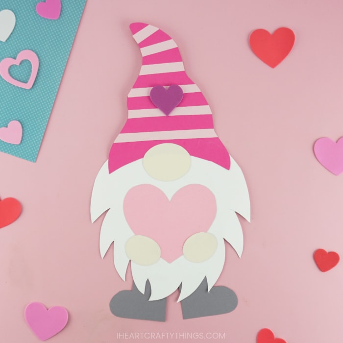 Gnome Valentine with a pink striped hat and gray shoes, holding a large pink paper heart.