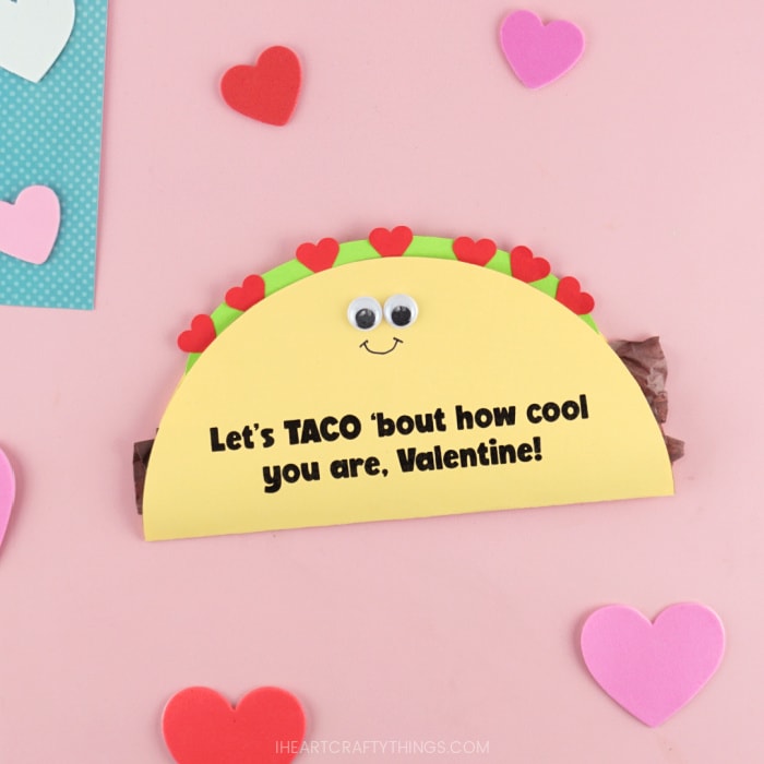 Close up image of DIY taco Valentine's Day card laying on a pink background with heart stickers scattered around.