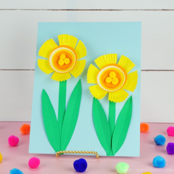 Cupcake liner Daffodil craft on a stand, sitting on a pink table with colored poms scattered around and a in front of a white shiplap background. 