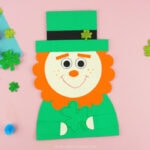 Completed leprechaun paper craft centered in the photo and laying on a pink background with glittery shamrock stickers and colored poms scattered around.