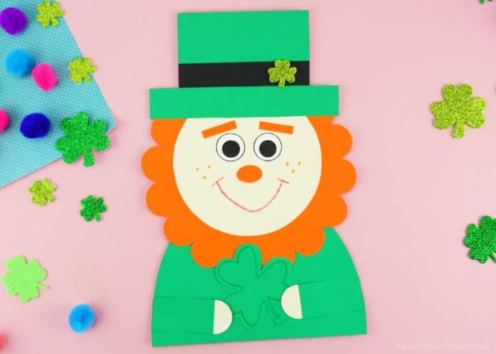 Horizontal image showing the finished leprechaun craft in the center of the photo laying on a pink background with glittery shamrock stickers and colored poms scattered around.