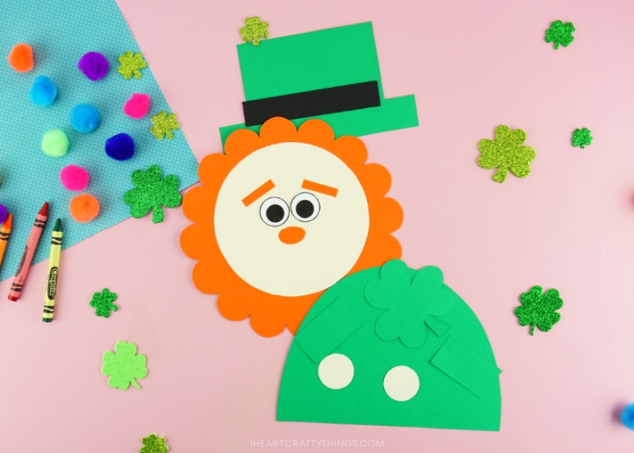 Each of the pieces from the leprechaun craft template cut out and laying on a pink background with shamrock stickers, colored poms and a few crayons scattered around them.