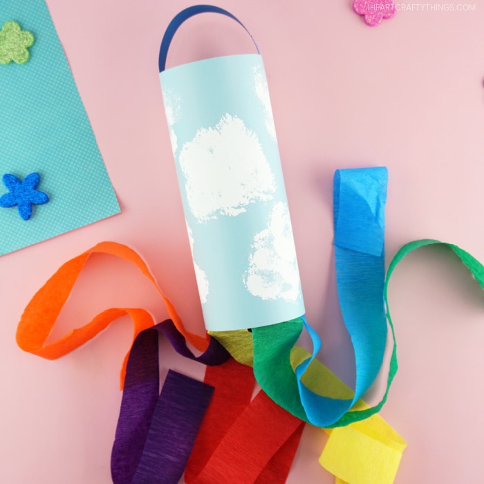 A kid-made rainbow windsock craft laying down flat on a pink background with the rainbow strips flowing out of the bottom of it in different directions.