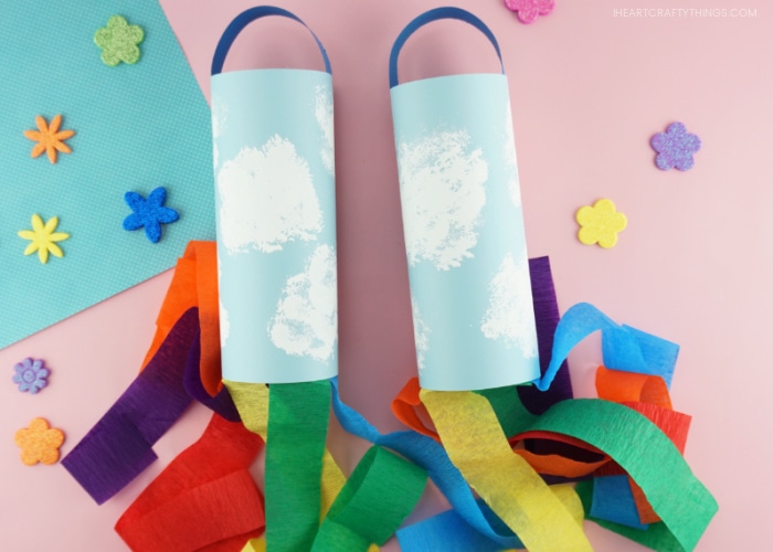 Two kid-made rainbow windsocks laying side by side on a pink background with flower stickers scattered around them.